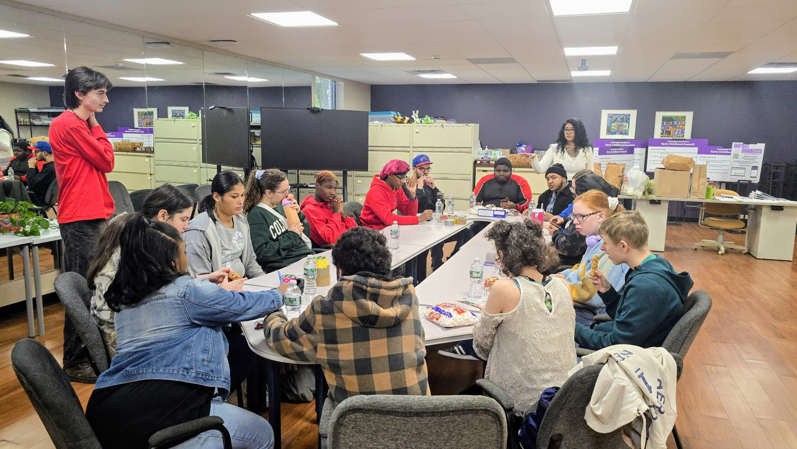 Students sitting at a oval shaped table with some drinking water and others have food in front of them