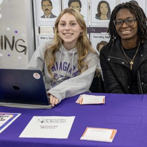 Students sitting at a table with laptops and sign for Community Service Hours